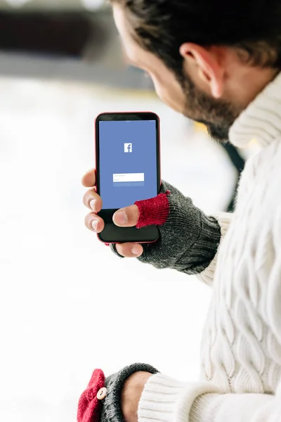 KYIV, UCRANIA - 15 de noviembre de 2019: vista recortada del hombre con guantes sosteniendo el teléfono inteligente con la aplicación de Facebook en la pantalla, en la pista de patinaje - foto de stock