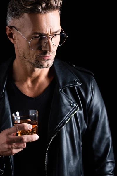Hombre guapo en chaqueta de cuero sosteniendo vaso de whisky aislado en negro - foto de stock