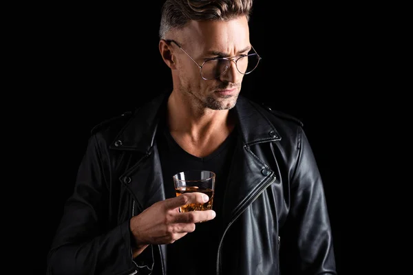 Stylish man in biker jacket and sunglasses holding glass of whiskey isolated on black — Stock Photo