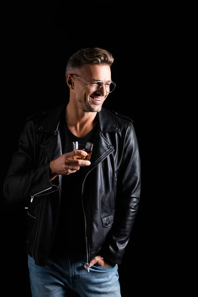 Smiling handsome man in leather jacket holding glass of whiskey isolated on black — Stock Photo