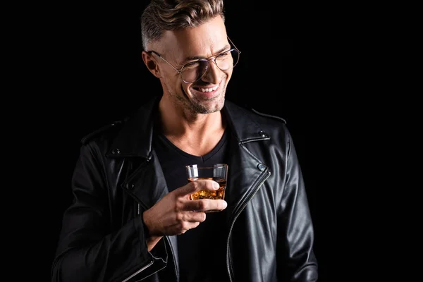 Hombre sonriente en chaqueta de cuero y gafas de sol sosteniendo vaso de whisky aislado en negro - foto de stock