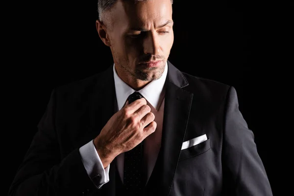 Gutaussehender Geschäftsmann passt Krawatte an, während er isoliert auf Schwarz wegschaut — Stockfoto