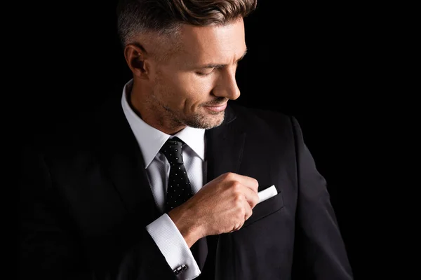 Hombre de negocios guapo en traje ajustando pañuelo aislado en negro - foto de stock