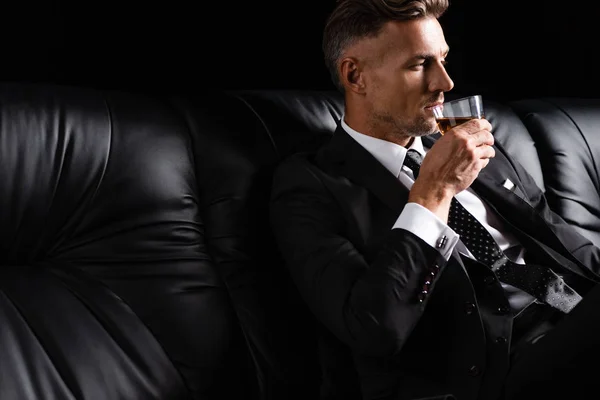 Hombre de negocios guapo bebiendo whisky mientras está sentado en el sofá aislado en negro — Stock Photo