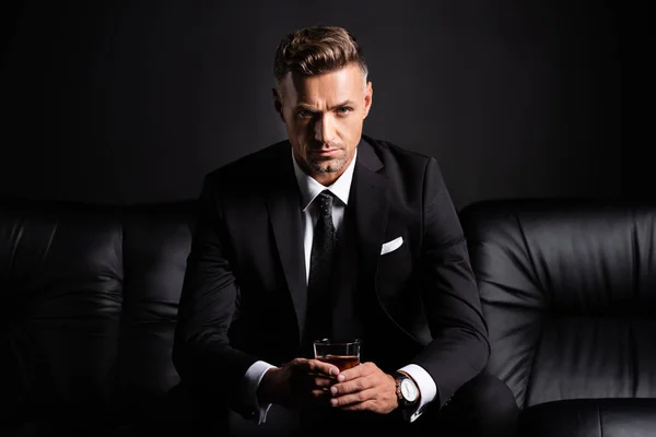 Hombre de negocios guapo con vaso de whisky mirando a la cámara en el sofá aislado en negro — Stock Photo