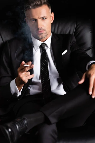Confident businessman looking at camera and smoking cigarette on sofa isolated on black — Stock Photo