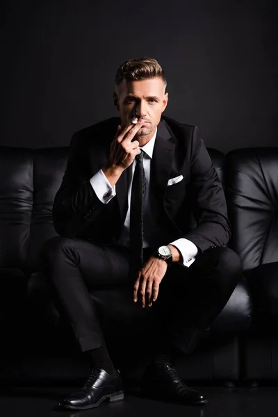 Businessman looking at camera while smoking cigarette on couch isolated on black — Stock Photo