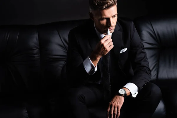 KYIV, UCRANIA - 11 DE OCTUBRE DE 2019: Empresario fumando iqos sentado en sofá aislado ob negro - foto de stock