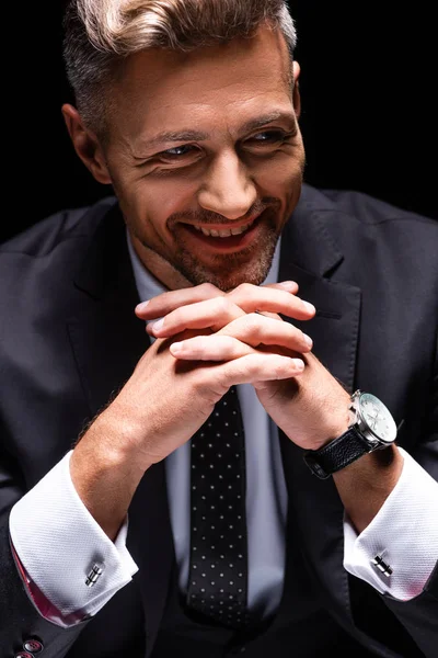 Hombre de negocios sonriente mirando hacia otro lado mientras cruza los dedos aislados en negro - foto de stock