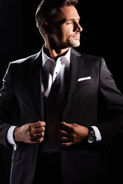 Hombre de negocios guapo ajustando solapas de chaqueta y mirando hacia otro lado aislado en negro - foto de stock