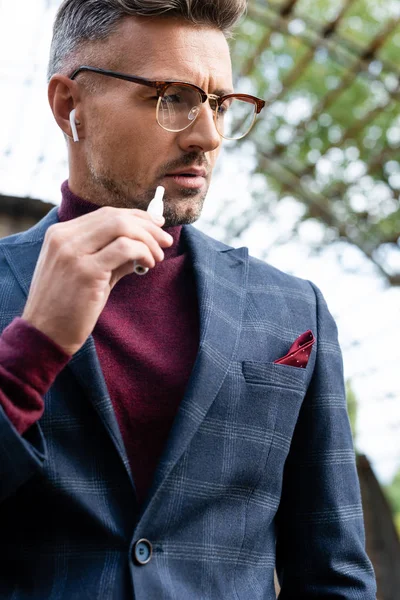 Handsome businessman in wireless earphones holding electronic cigarette outdoors — Stock Photo