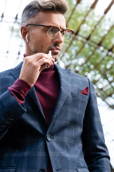 Vista de bajo ángulo del hombre de negocios en auriculares inalámbricos fumar cigarrillo electrónico al aire libre — Stock Photo