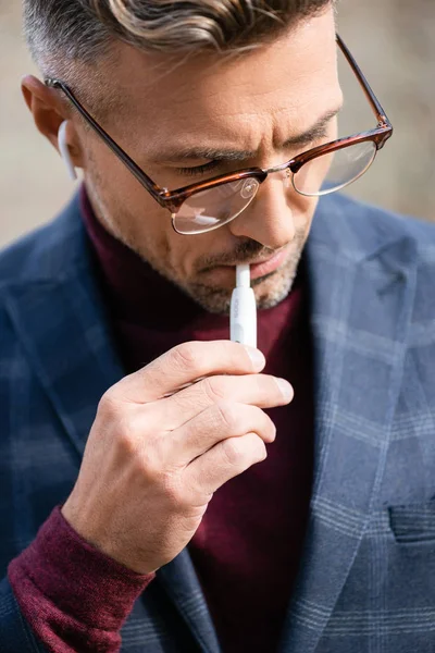 KYIV, UCRANIA - 11 de octubre de 2019: Elegante hombre de negocios con gafas y auriculares inalámbricos fumando iqos — Stock Photo