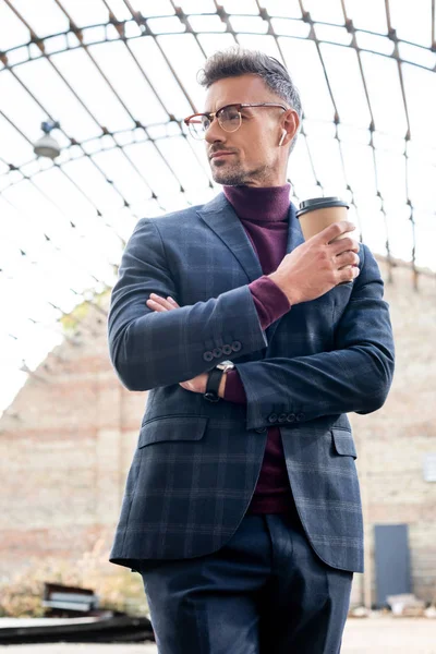 Businessman in wireless earphones holding disposable cup on urban street — Stock Photo