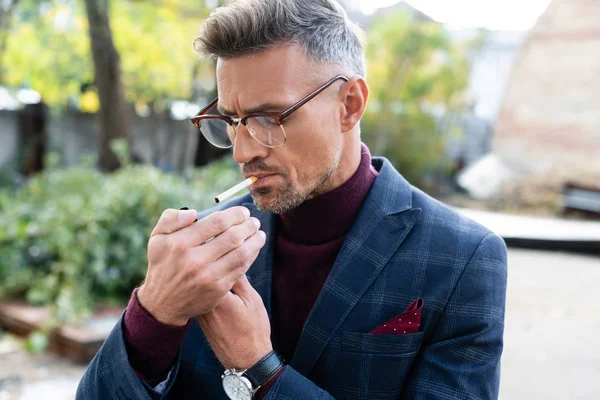 Handsome businessman in eyeglasses lighting cigarette on urban street — Stock Photo