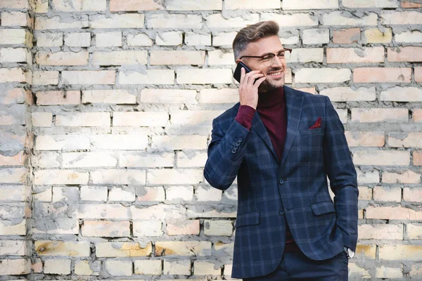 Smiling businessman talking on smartphone with brick wall at background — Stock Photo