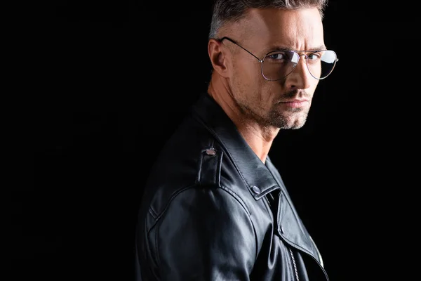 Side view of serious man in sunglasses and leather jacket looking at camera isolated on black — Stock Photo