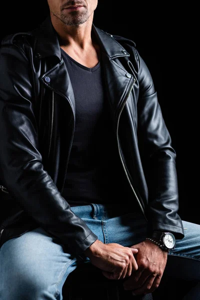 Обрезанный вид стильного человека в кожаной куртке и джинсах, сидящих на стуле изолированы на черном — стоковое фото