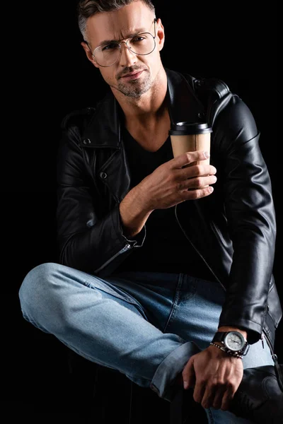 Beau homme en veste en cuir avec café pour aller regarder la caméra isolée sur noir — Photo de stock