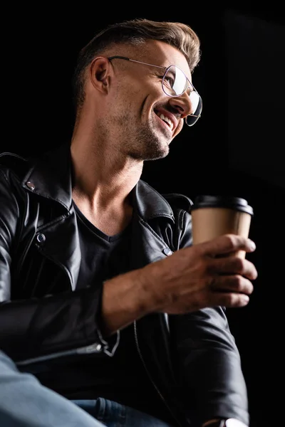 Vista de ángulo bajo del hombre sonriente en gafas de sol y chaqueta de cuero que sostiene el café para ir aislado en negro - foto de stock