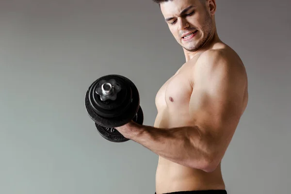 Боковой вид сексуального мускулистого культуриста с голым туловищем, тренирующимся с гантели, изолированной на сером — стоковое фото