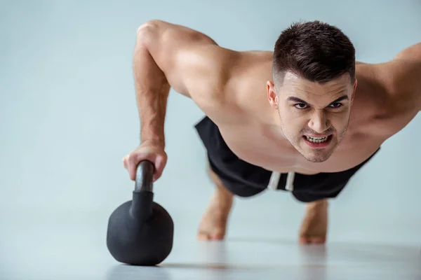 Sexy muscular bodybuilder with bare torso doing push ups with kettlebell on grey — Stock Photo