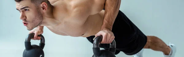 Sexy muscular bodybuilder with bare torso doing push ups with kettlebells isolated on grey, panoramic shot — Stock Photo