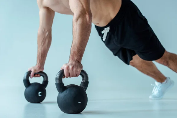 Partial view of sexy muscular bodybuilder with bare torso doing push ups with kettlebells on grey — Stock Photo