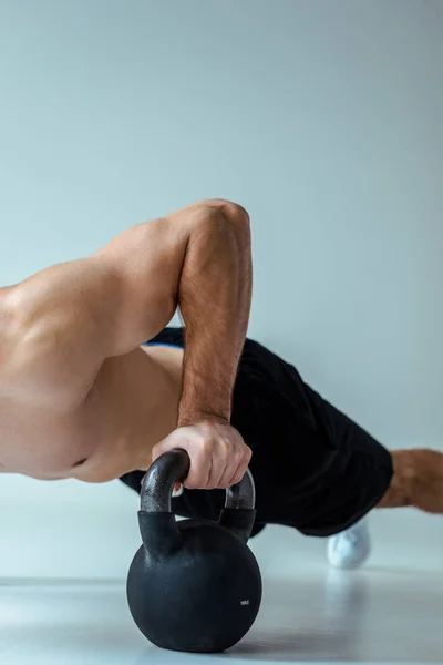 Partial view of sexy muscular bodybuilder with bare torso doing push ups with kettlebell on grey — Stock Photo