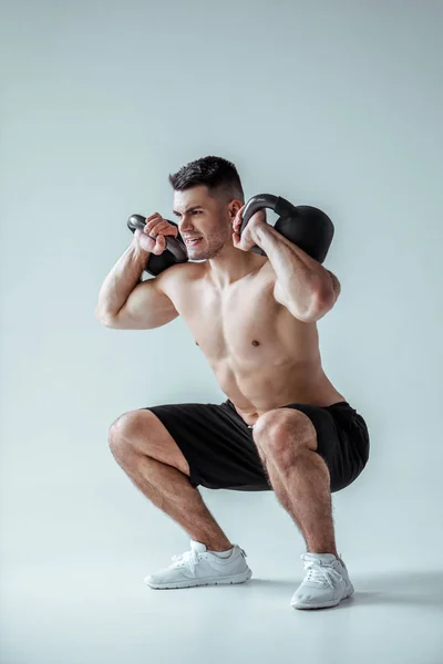 Sexy muscular bodybuilder with bare torso squatting with kettlebells on grey — Stock Photo