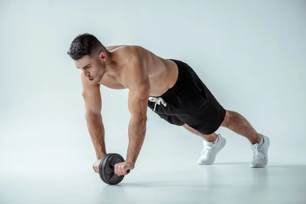 Sexy muscular bodybuilder with bare torso exercising with ab wheel on grey background — Stock Photo