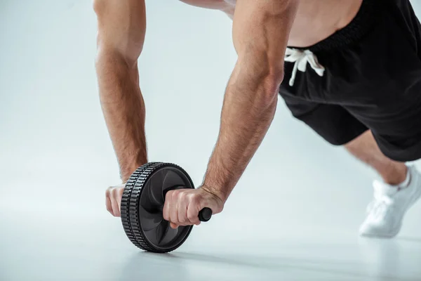 Vista recortada de culturista muscular sexy con torso desnudo haciendo ejercicio con rueda abdominales sobre fondo gris - foto de stock