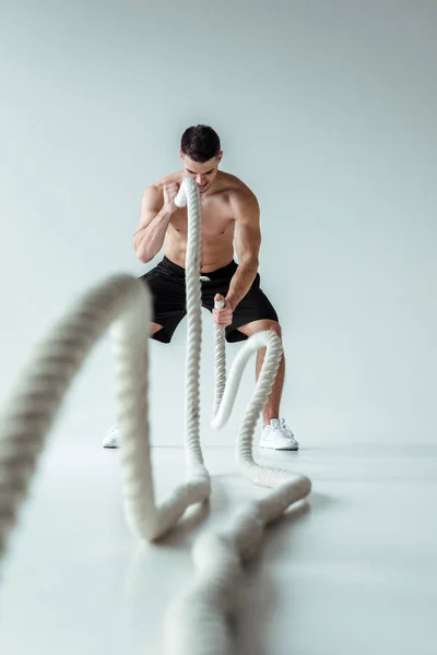 Selective focus of sexy muscular bodybuilder with bare torso exercising with battle rope on grey background — Stock Photo