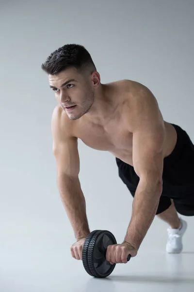Sexy musculoso culturista con torso desnudo ejercitando con rueda abdominales sobre fondo gris - foto de stock