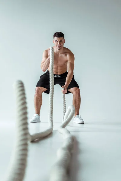 Foco seletivo do fisiculturista muscular sexy com tronco nu exercitando com corda de batalha no fundo cinza — Fotografia de Stock