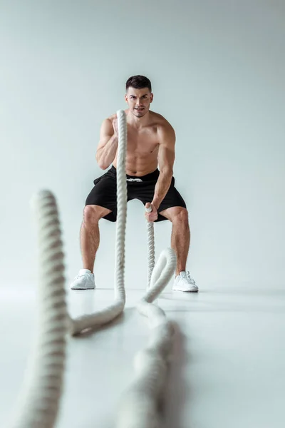 Selective focus of sexy muscular bodybuilder with bare torso exercising with battle rope on grey background — Stock Photo