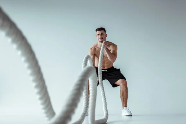 Selective focus of sexy muscular bodybuilder with bare torso exercising with battle rope on grey background — Stock Photo