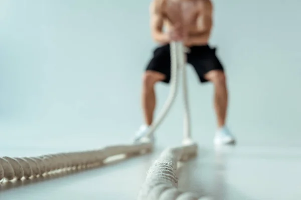 Selective focus of sexy muscular bodybuilder with bare torso exercising with battle rope on grey background — Stock Photo