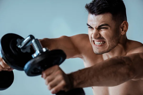 Selective focus of tense muscular bodybuilder with bare torso working out with dumbbells isolated on grey — Stock Photo