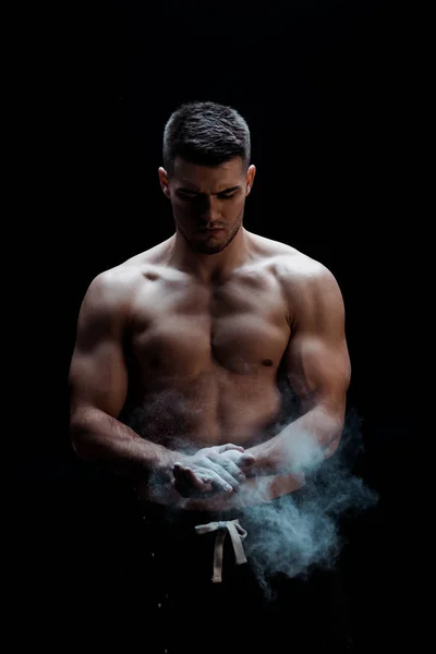 Sexy muscular bodybuilder with bare torso applying talcum powder on hands on black background with smoke — Stock Photo