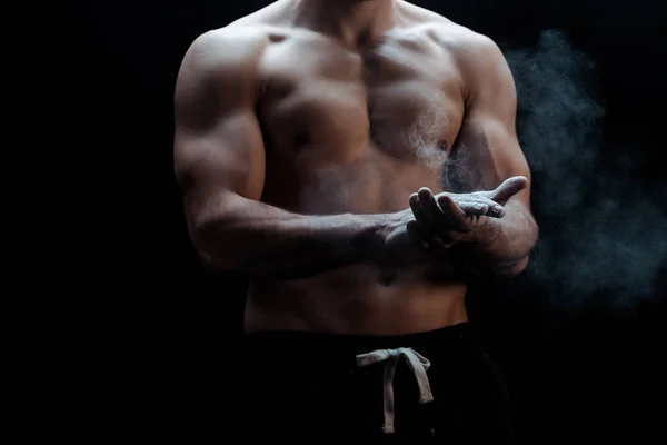 Cropped view of sexy muscular bodybuilder with bare torso applying talcum powder on hands on black background with smoke — Stock Photo