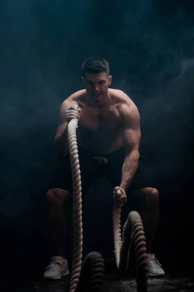 Sexy muscular bodybuilder with bare torso excising with battle rope on black background with smoke — Stock Photo
