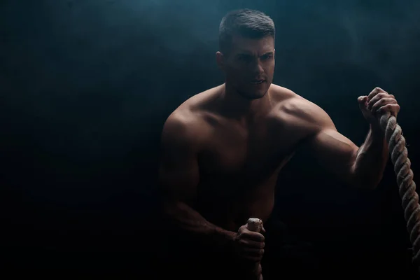 Sexy muscular bodybuilder with bare torso excising with battle rope on black background with smoke — Stock Photo