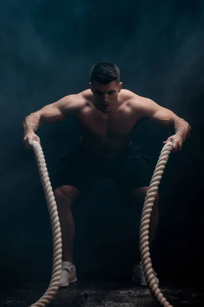Сексуальний м'язистий культурист з голим торсом займається з бойовою мотузкою на чорному тлі з димом — стокове фото