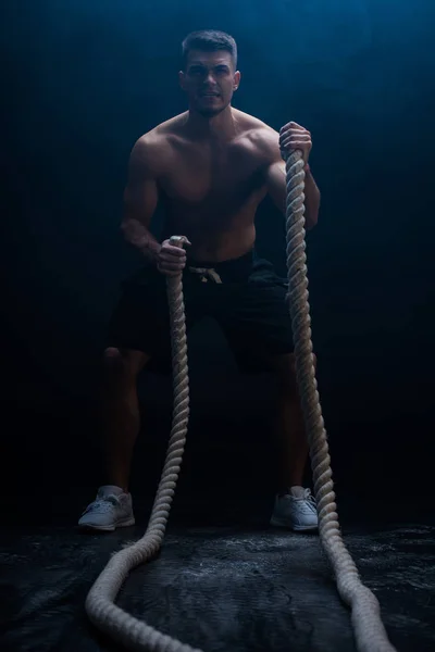 Selektiver Fokus des sexy muskulösen Bodybuilders mit nacktem Oberkörper beim Schneiden mit dem Kampfseil auf schwarzem Hintergrund mit Rauch — Stockfoto