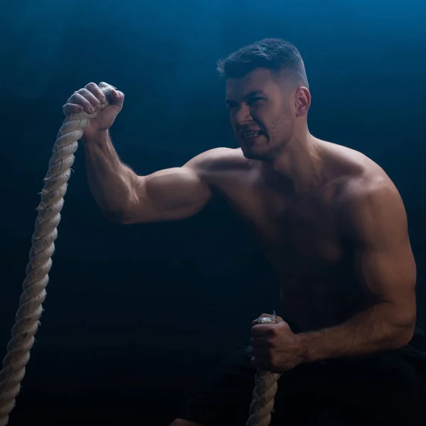 Tense muscular bodybuilder with bare torso excising with battle rope on black background with smoke — Stock Photo