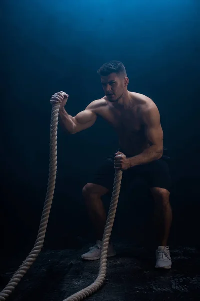 Sexy muscular bodybuilder with bare torso excising with battle rope on black background with smoke — Stock Photo