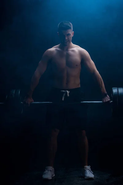 Sexy muscular bodybuilder with bare torso excising with barbell on black background with smoke — Stock Photo