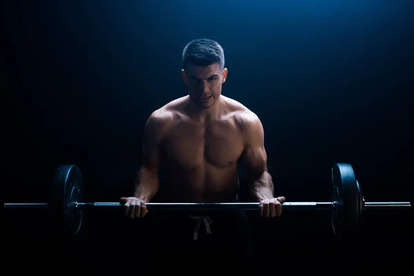 Sexy muscular bodybuilder with bare torso excising with barbell on black background with smoke — Stock Photo