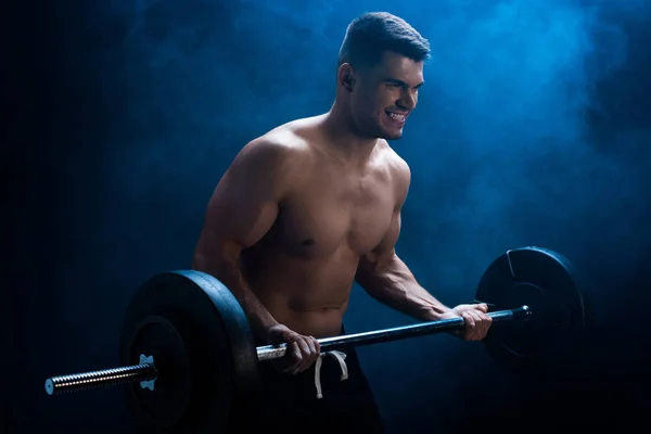 Fisiculturista muscular sexy com tronco nu extirpando com barbell no fundo preto com fumaça — Fotografia de Stock